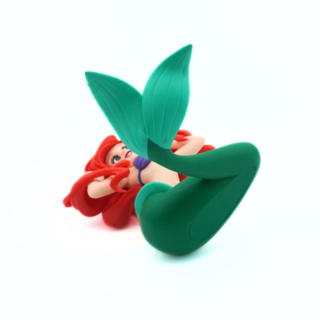 A small mermaid figurine or plush toy with bright red hair and a green tail, posed on its side against a plain white background.