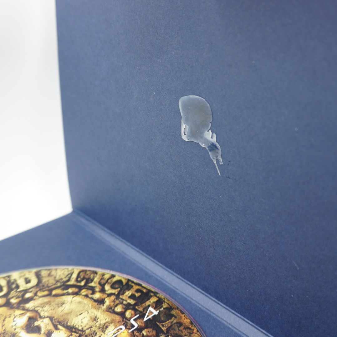A close-up of a small clear, dome-shaped droplet or bubble resting on the inside of a dark blue box, with part of a gold-toned circular object visible at the bottom left.