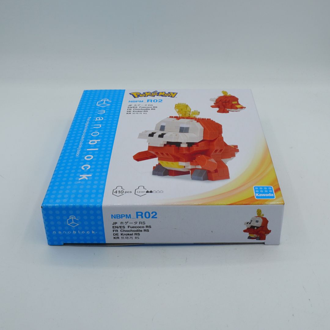 A boxed nanoblock toy set for Pokémon, specifically Fuecoco, shown on a plain light gray studio background. The retail box is white and blue with a large photo of the assembled red-and-white dragon-like toy on the front and smaller product details printed on the sides.