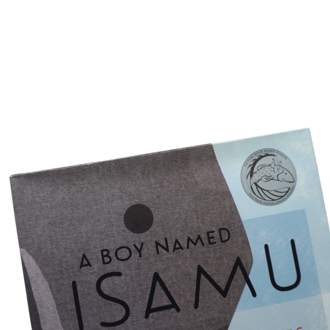 A close-up of a printed card or box cover with the text 'A BOY NAMED ISAMU' on a dark gray and pale blue background, plus a circular fish logo.