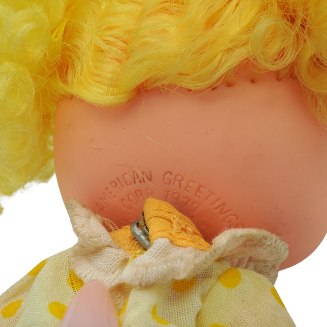 Close-up of a doll with yellow hair and a yellow dress with polka dots, featuring the American Greetings logo.
