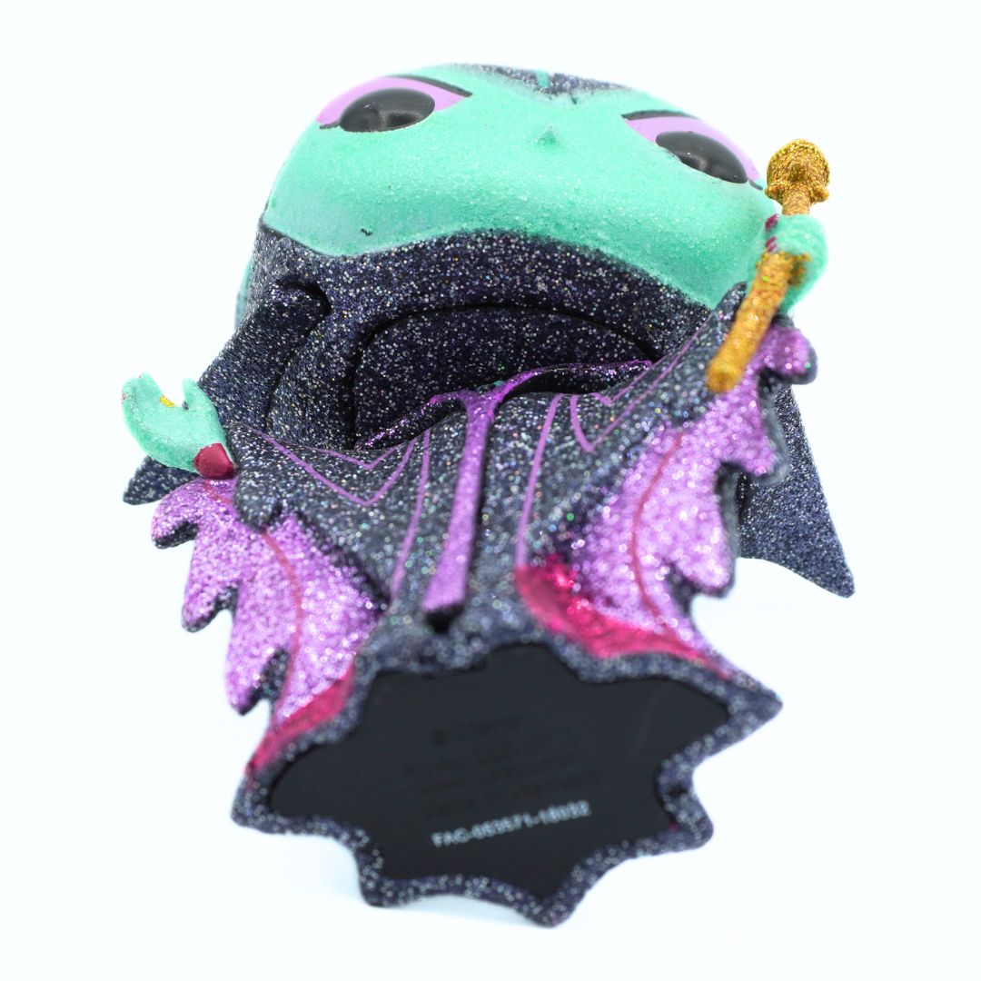 A collectible figurine of a green-skinned character, featuring a black glittery cape with purple and pink glitter accents, a gold staff, and a mint green top, shown against a plain white background.