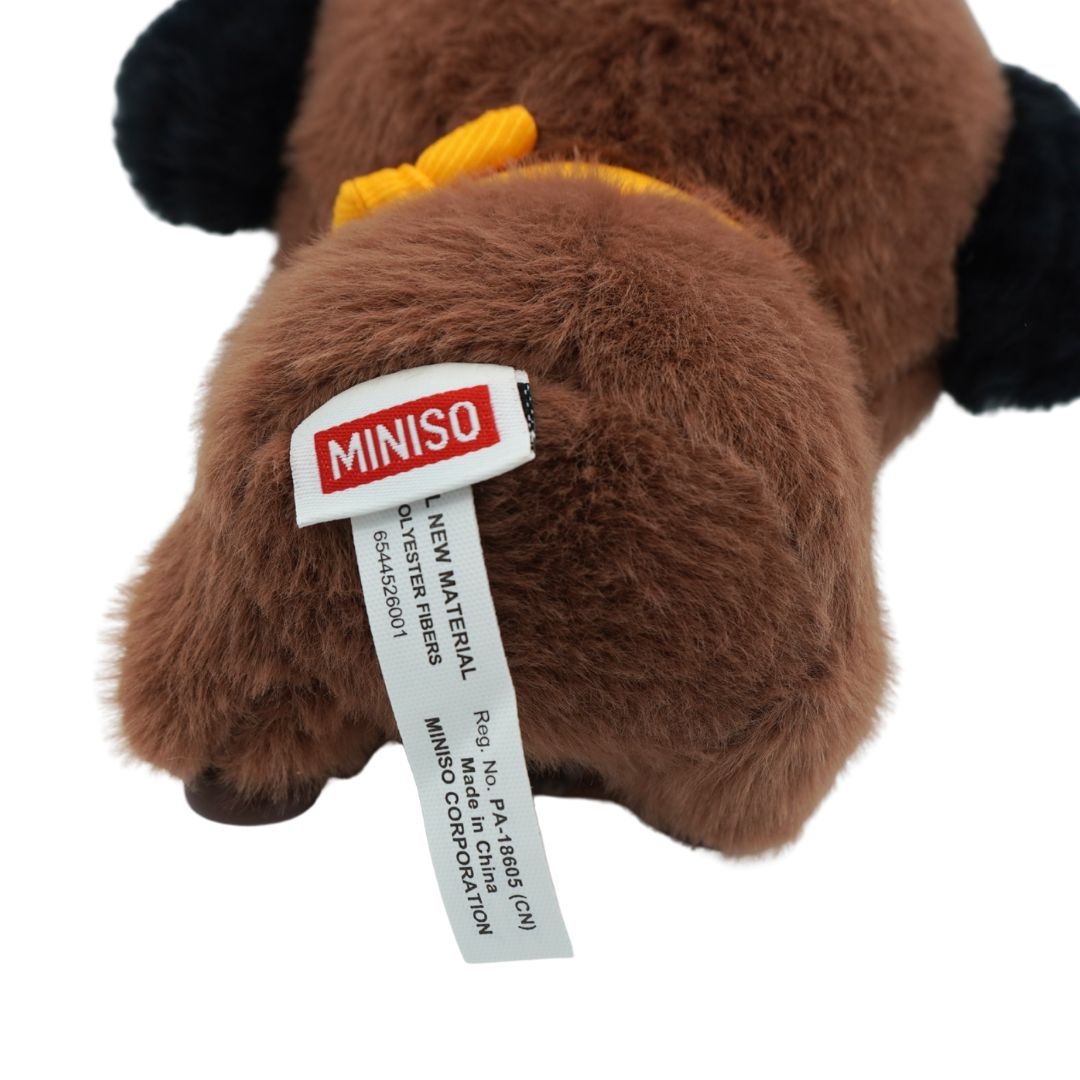 Close-up of a brown plush toy with black ears and a yellow bow, showing a sewn-on MINISO tag on a white background.
