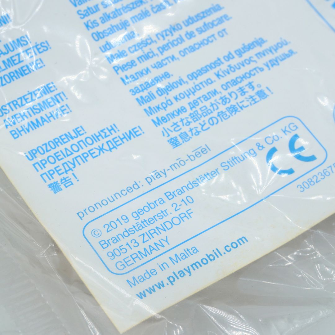 Close-up of the back of a white plastic product package showing printed blue text, warning labels, a CE mark, and the brand name play-mobil.
