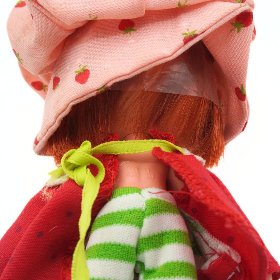 Close-up of a doll with red hair, wearing a pink hat with strawberry pattern, red coat, and green striped socks.