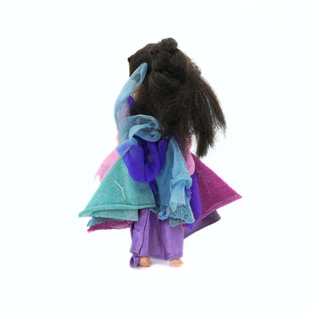 A vintage doll viewed from the back, featuring long dark brown hair and a layered, multi-colored dress in shades of purple, teal, pink, and blue, set against a plain white background.