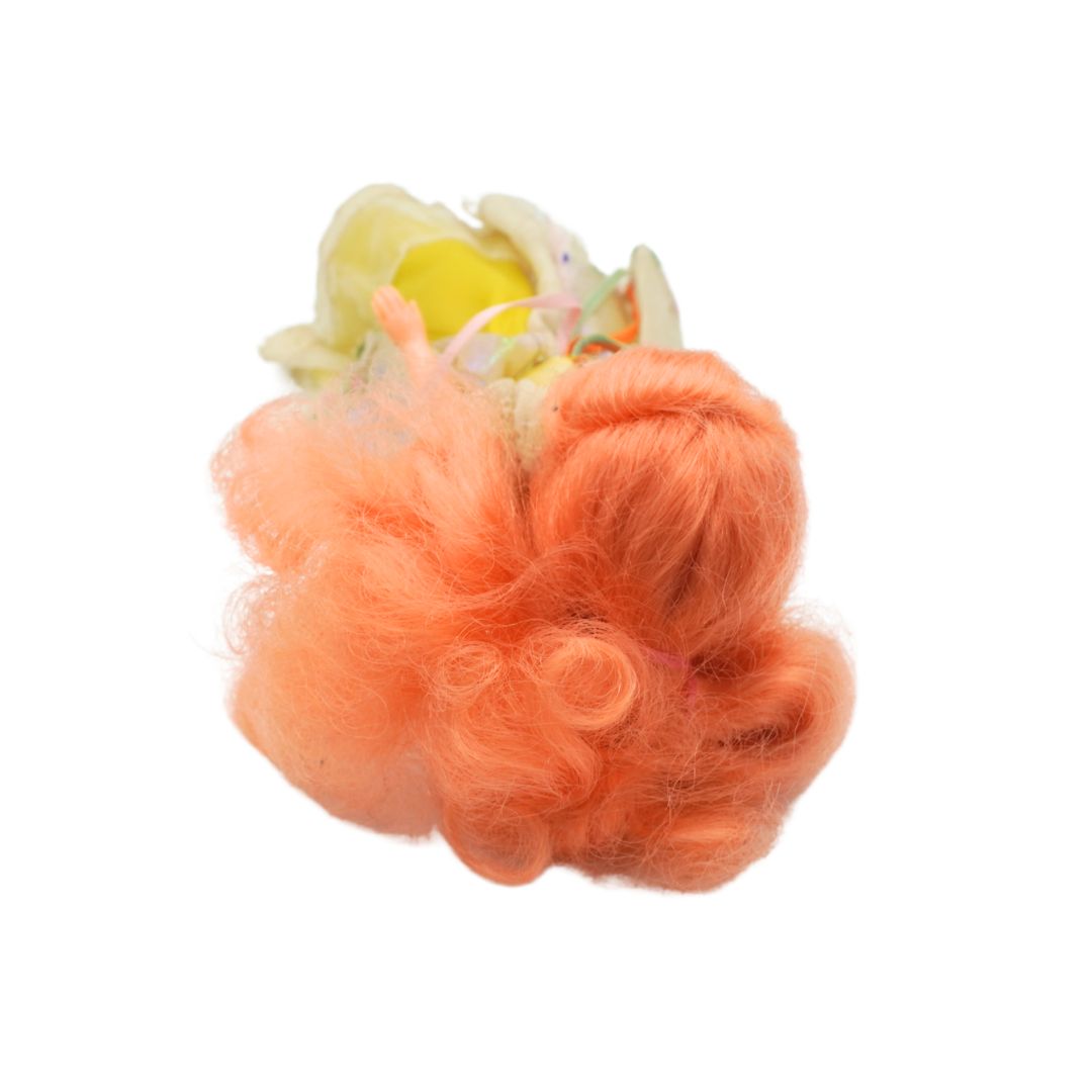 A fluffy, curly orange synthetic hairpiece or wig topper with a small pale yellow and white flower accent at the top, isolated on a white background.