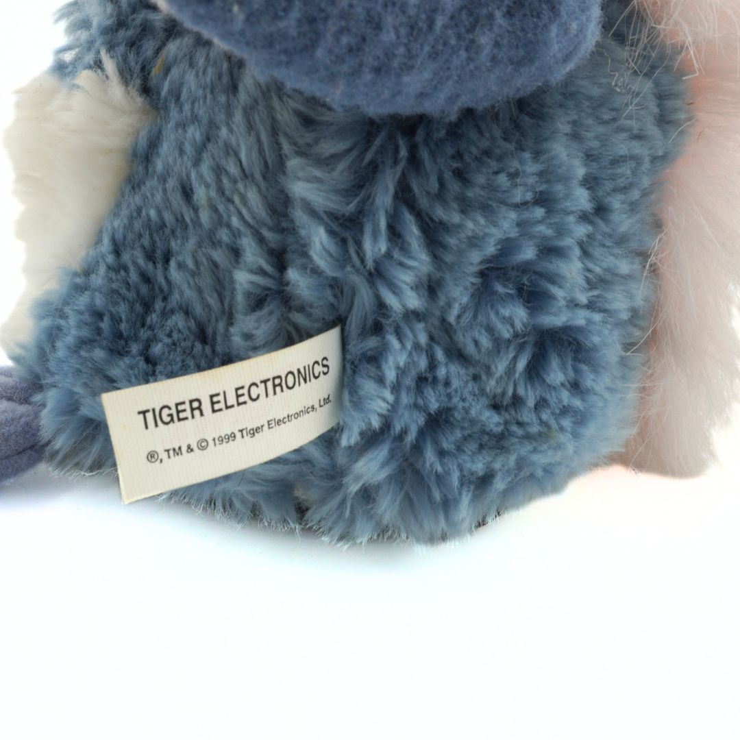 Close-up of a blue plush toy with a Tiger Electronics label on a white background