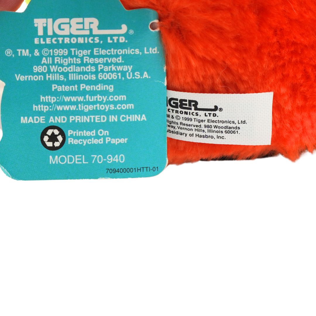 An orange and green Furby with yellow hair and blue eyes, photographed against a white background
