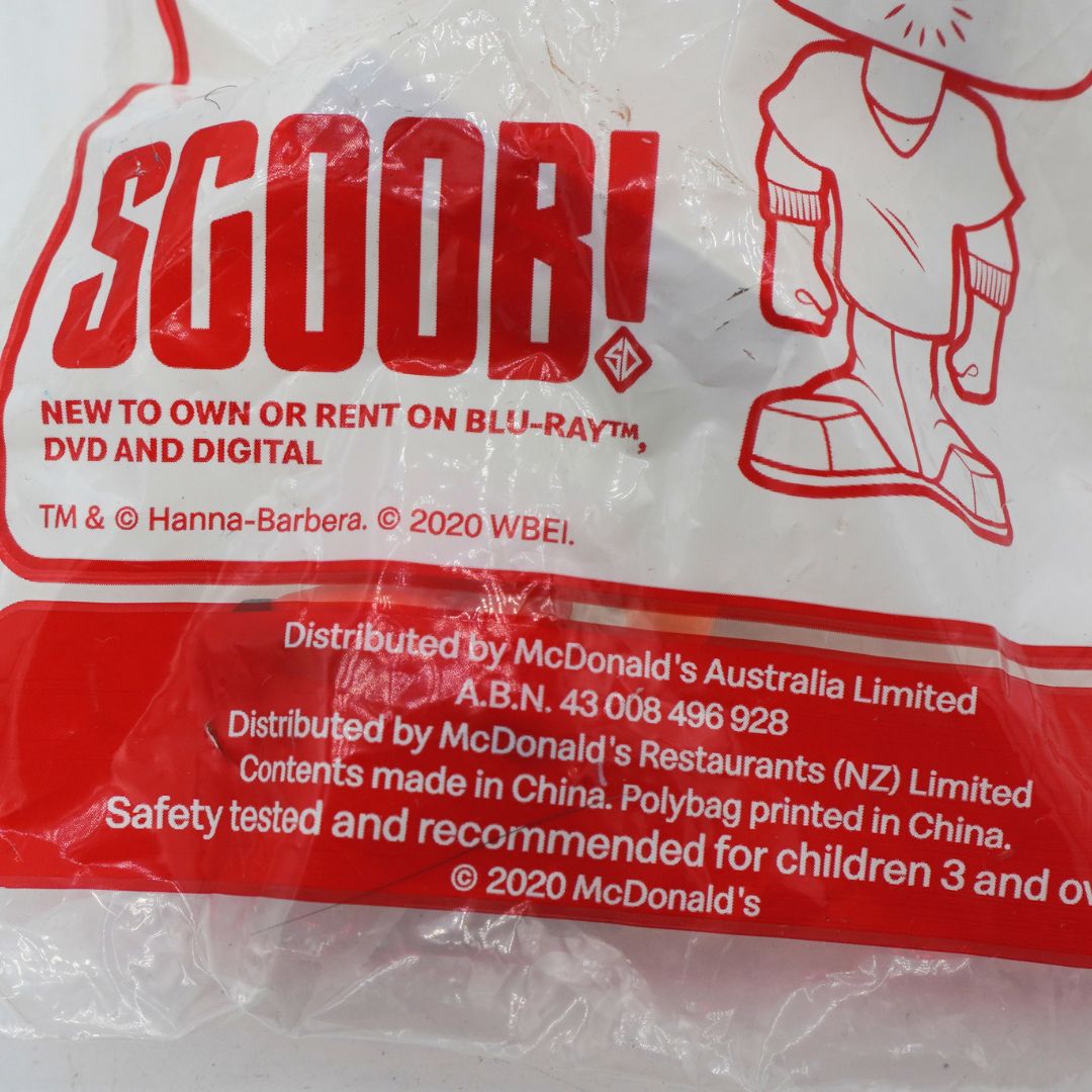 A close-up of a plastic bag containing a Scooby! branded item, with large red 'SCOOB!' text, a red outline illustration, and printed distribution information for McDonald's Australia and McDonald's Restaurants (NZ) Limited.