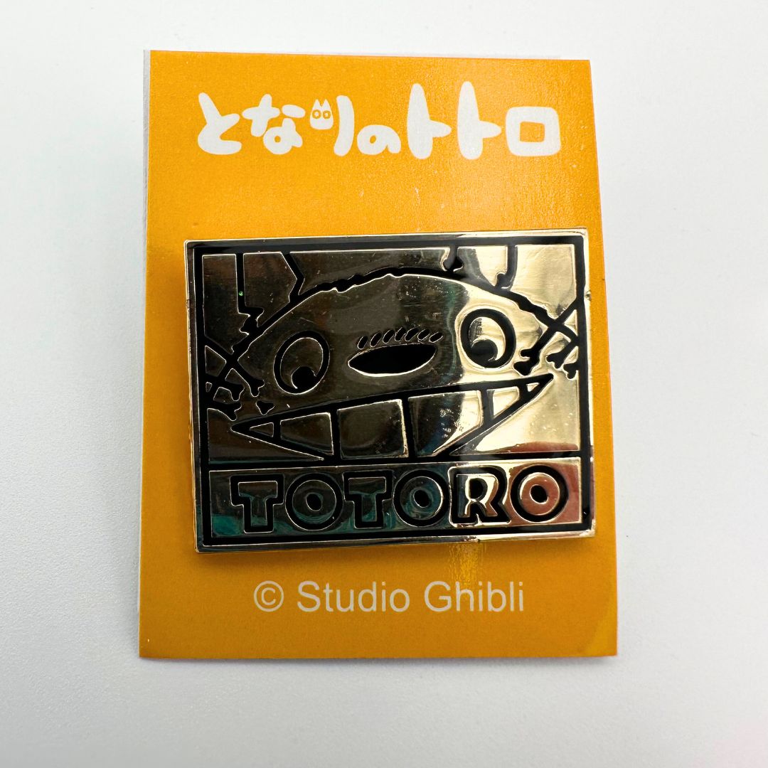 A front on photo of a gold-toned Studio Ghibli Totoro pin on an orange cardback