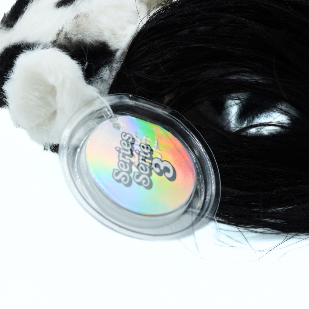 A close-up of a black and white cow-print plush toy with a shiny black synthetic wig and a round iridescent tag reading 'Series Series 3' on a white background.