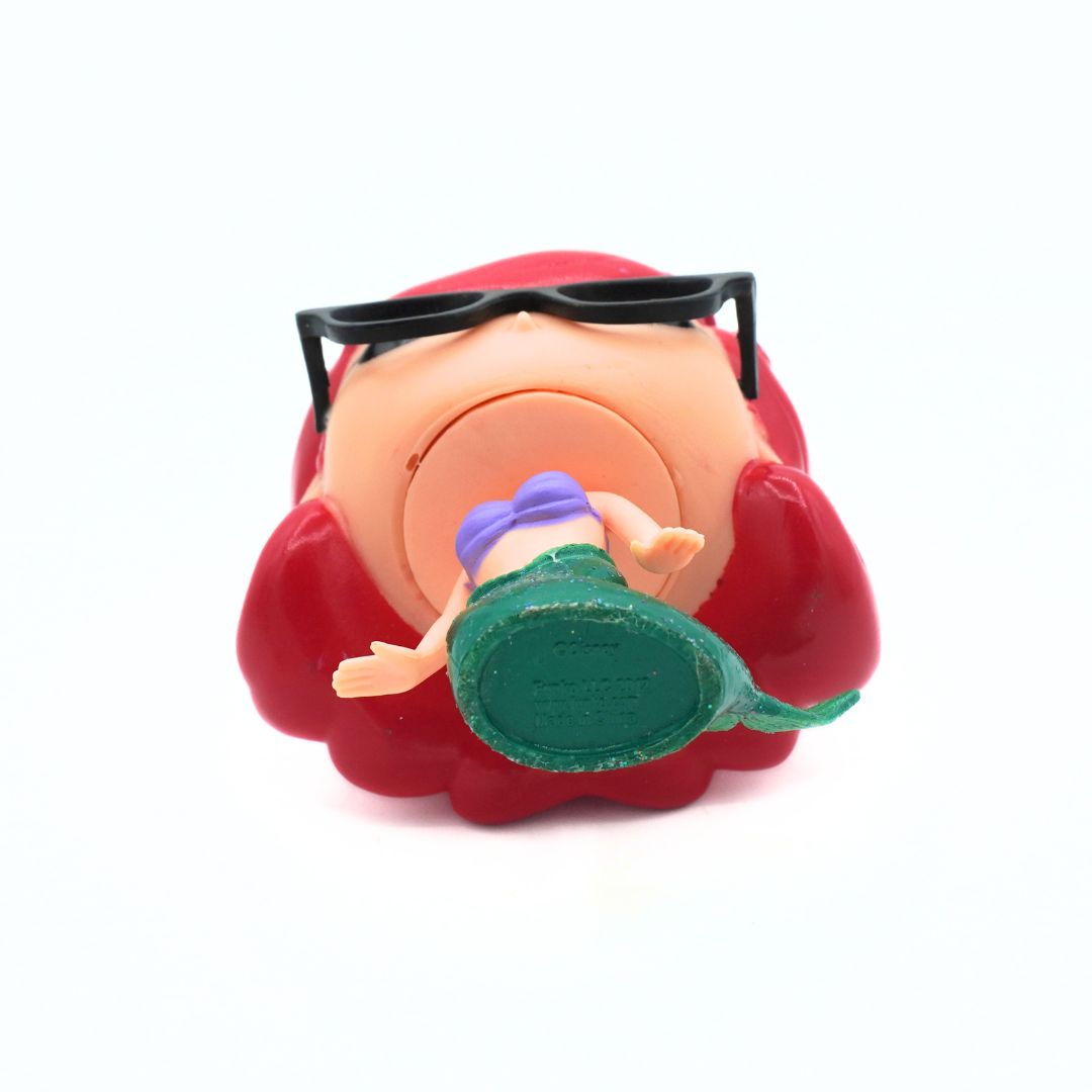 A vintage plastic toy figure of a woman with red hair and black sunglasses, viewed from the back to show the underside with a green base.