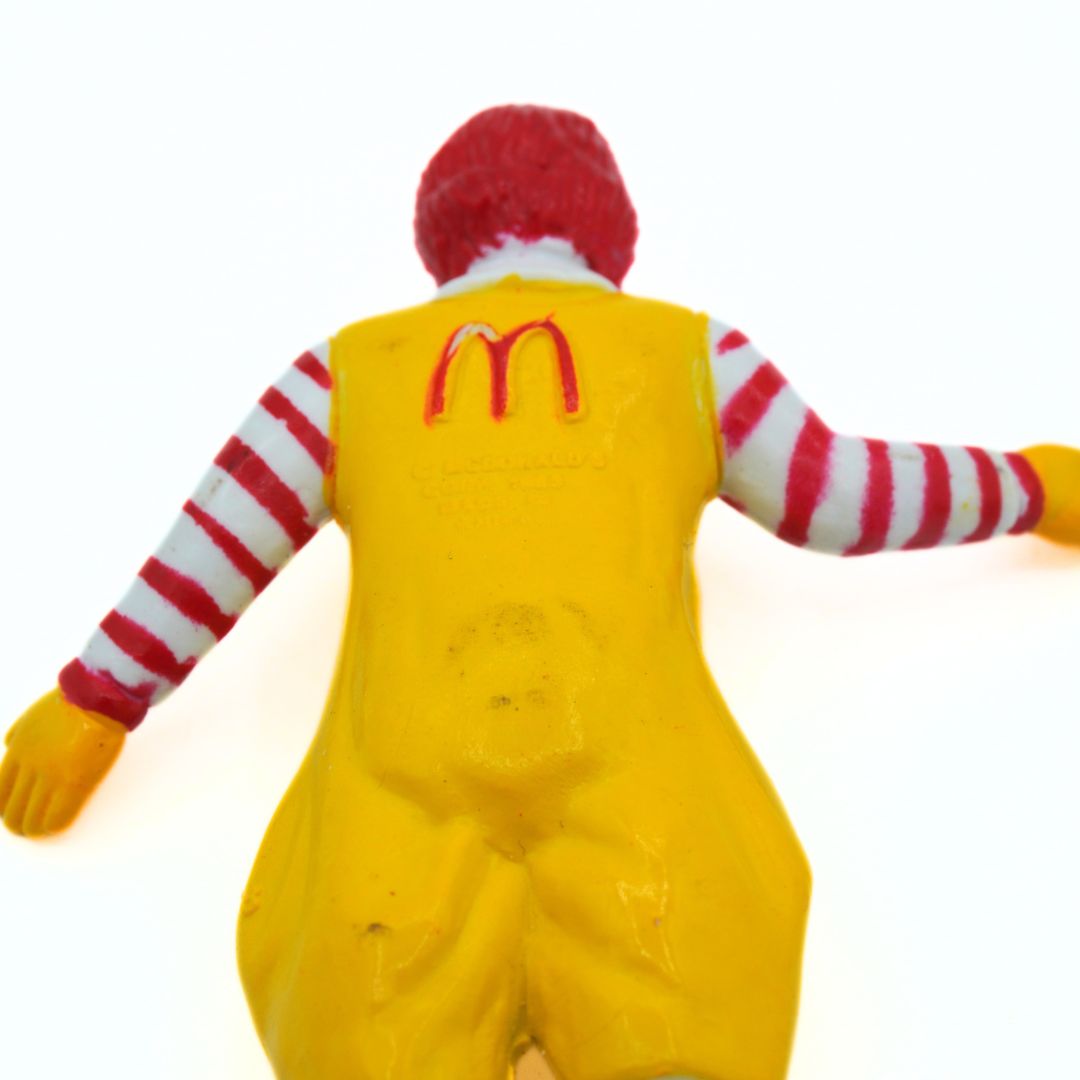 Back view of a Ronald McDonald action figure wearing a yellow jumpsuit, red and white striped shirt, and red hair against a plain white background.