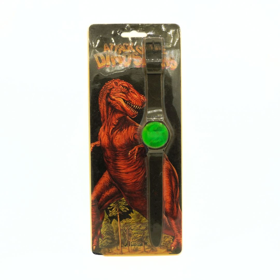 A black plastic dinosaur-shaped wristwatch with a bright green circular dial, presented in its original blister packaging featuring a red T-Rex illustration.