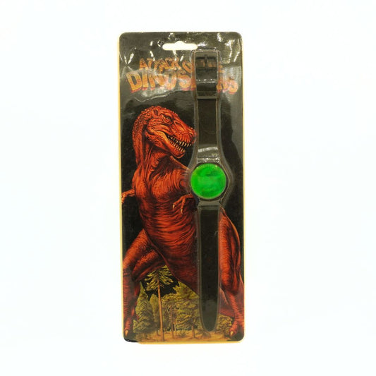 A black plastic dinosaur-shaped wristwatch with a bright green circular dial, presented in its original blister packaging featuring a red T-Rex illustration.