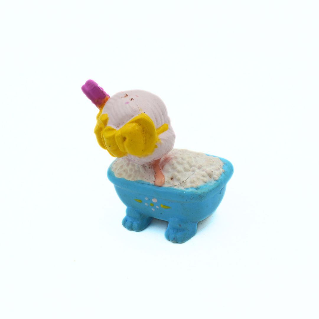 Toy figure of a character with a yellow bow and pink hair accessory sitting in a blue bathtub on a white background