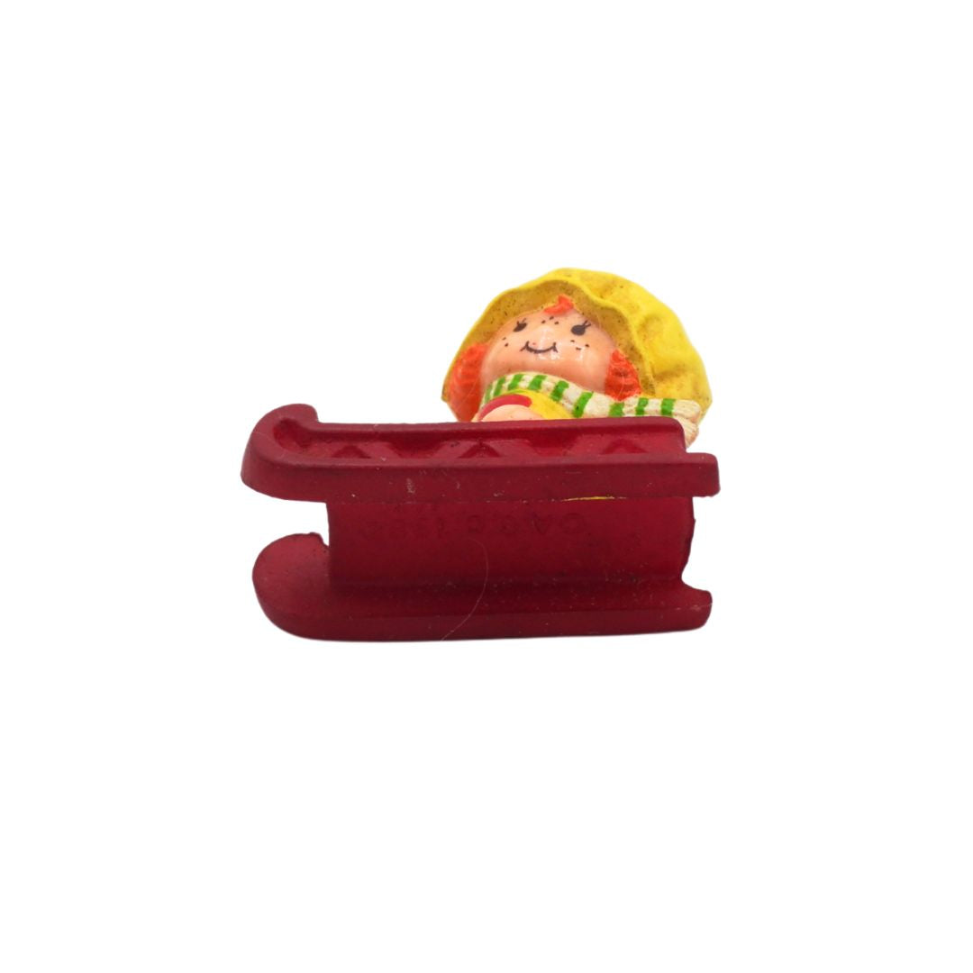 Small figurine of a person in a yellow raincoat sitting in a red sled on a white background