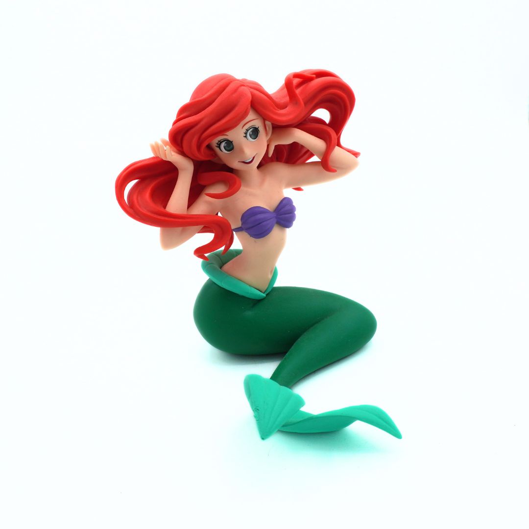 A small figurine of a mermaid with long red hair, a purple top, and a green tail, posed sitting against a plain white background.