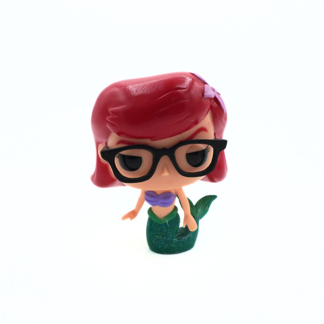 A small vinyl figurine of a mermaid girl with short red hair, black glasses, a purple top, and a green mermaid tail, standing against a plain white background.