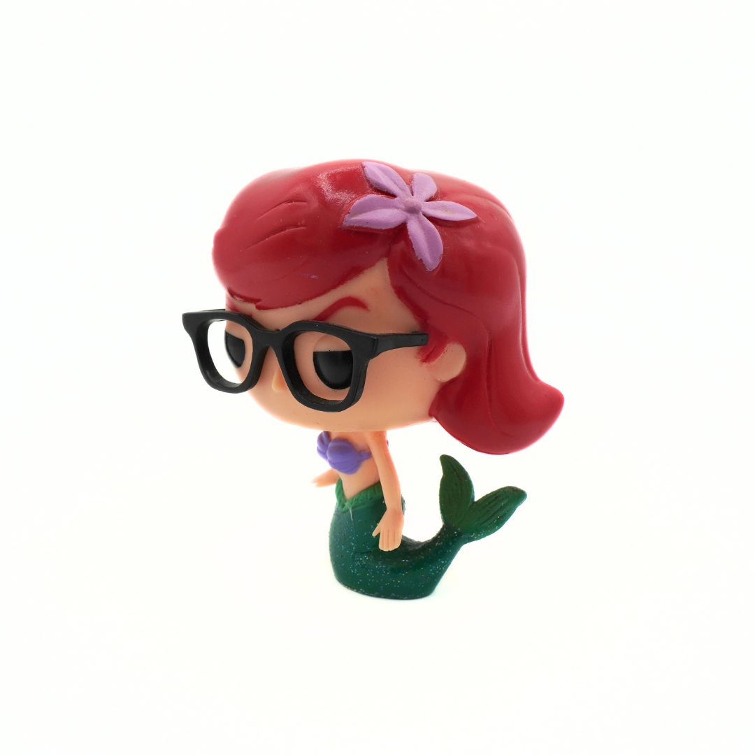 A small stylized mermaid figurine with red hair, a purple flower accessory, black glasses, and a green tail on a plain white background.