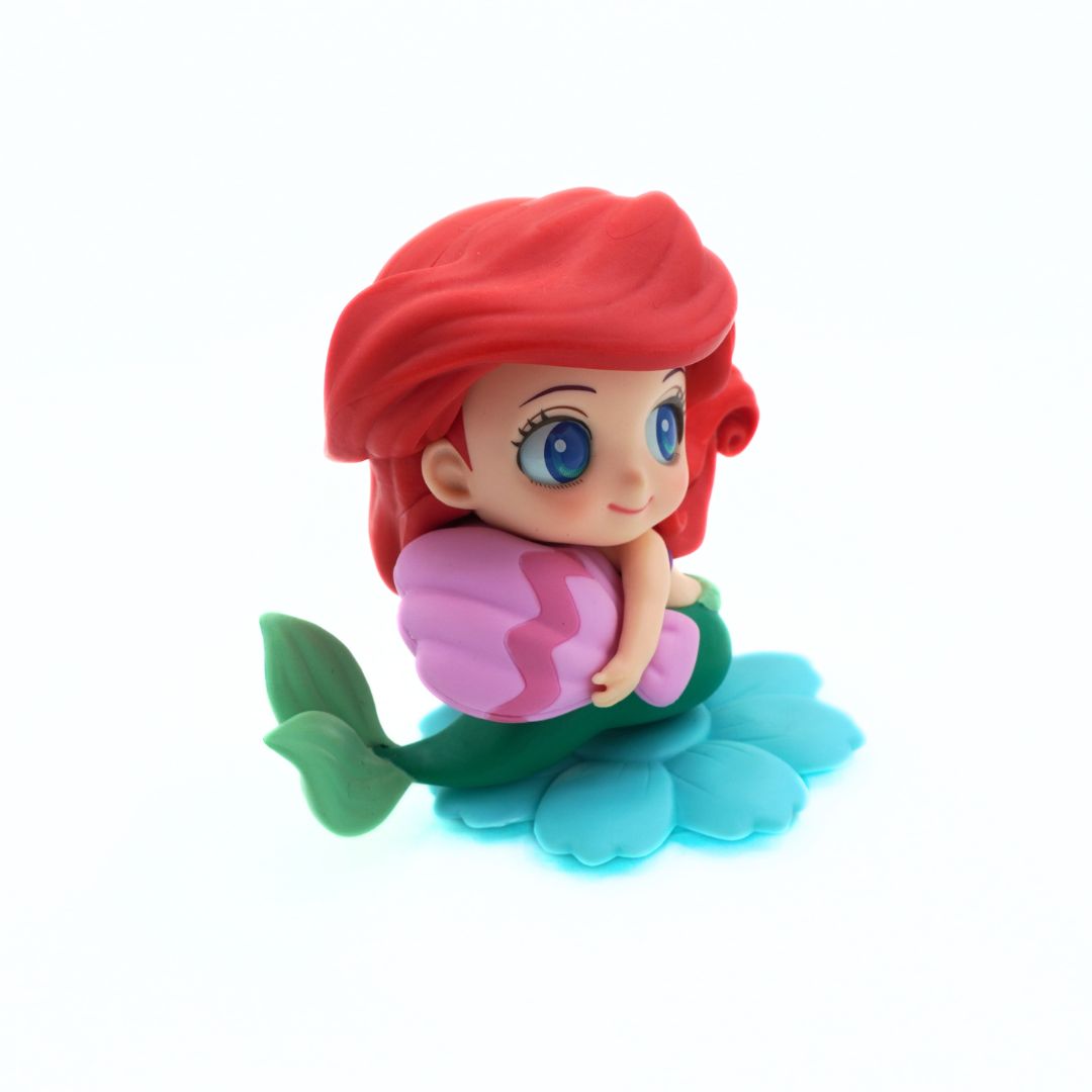 A small mermaid figurine with red hair, blue eyes, a green tail, and a pink shell, seated on a light blue flower-like base against a plain white background.