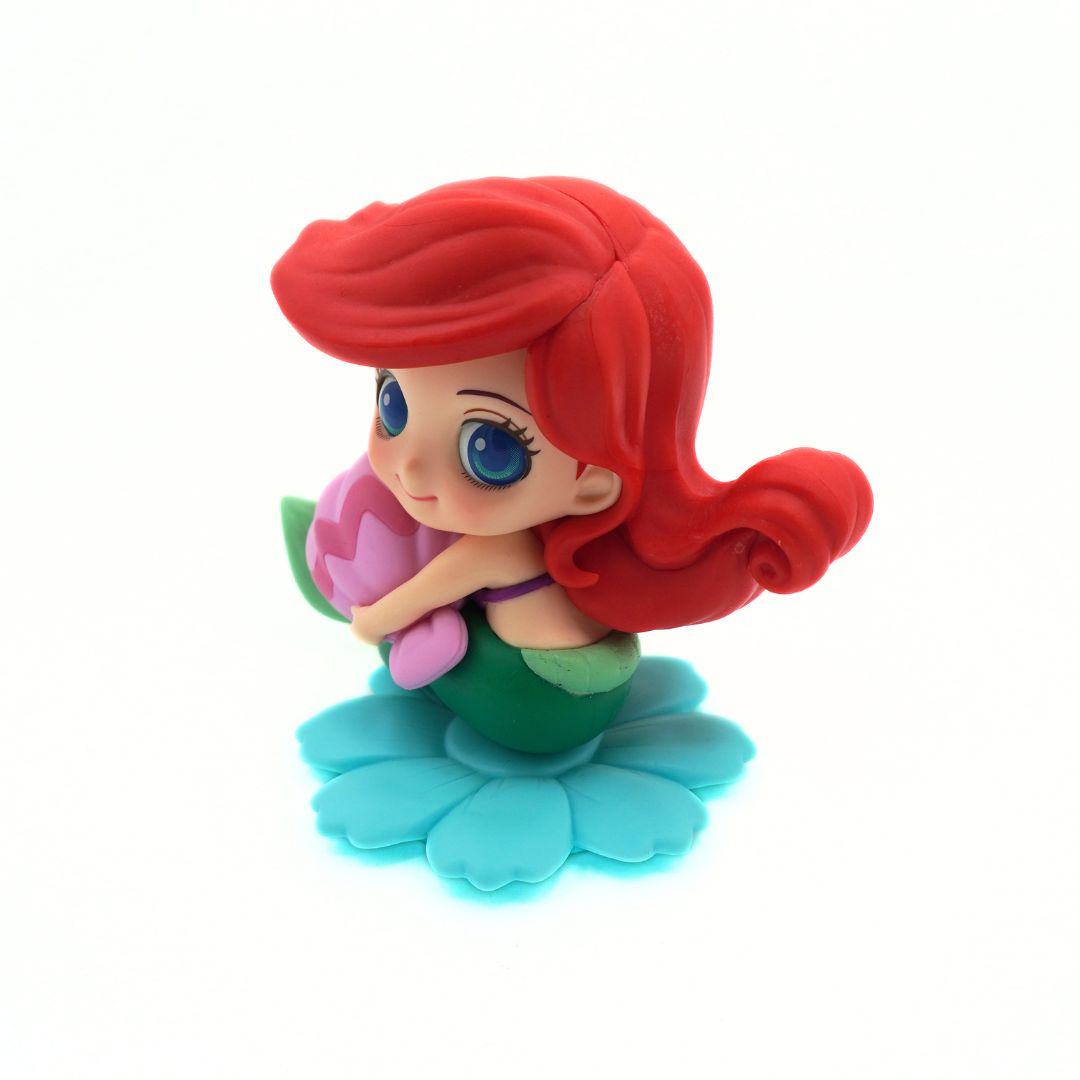 A small Ariel-themed figurine with long red hair, blue eyes, a green and purple top, holding a pink flower, seated on a turquoise flower-shaped base against a plain white background.