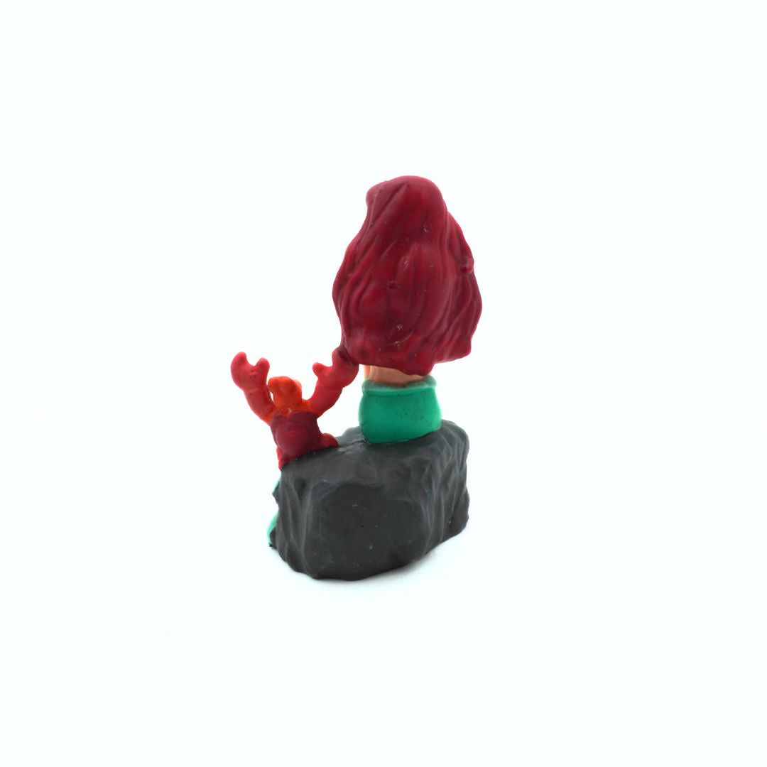 Small figurine of a character with red hair and green clothing sitting on a rock against a white background