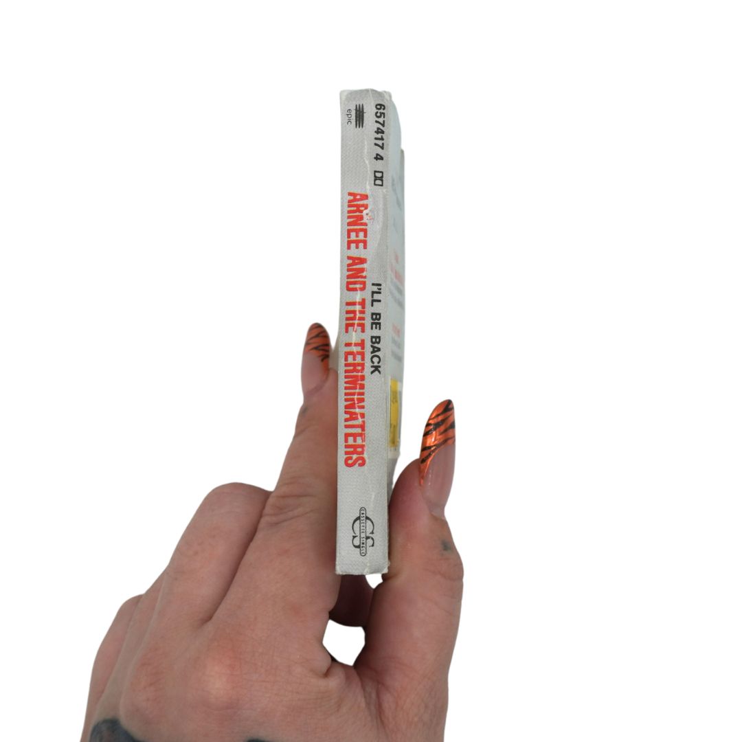 A hand with orange and black patterned nail art holds a slim white book or card featuring the title 'ARNEE AND THE TERMINATORS' in red text, against a plain white background.