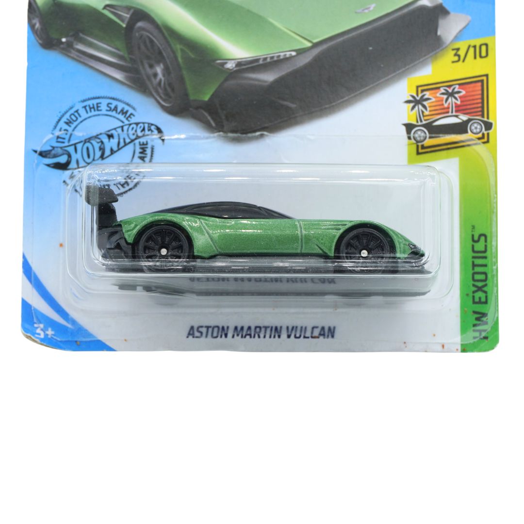A Hot Wheels die-cast model car of the Aston Martin Vulcan, shown in its original blister packaging. The toy car is metallic green with black wheels and a black rear spoiler, set against a blue and green card backing with the Hot Wheels logo and 'Aston Martin Vulcan' text.
