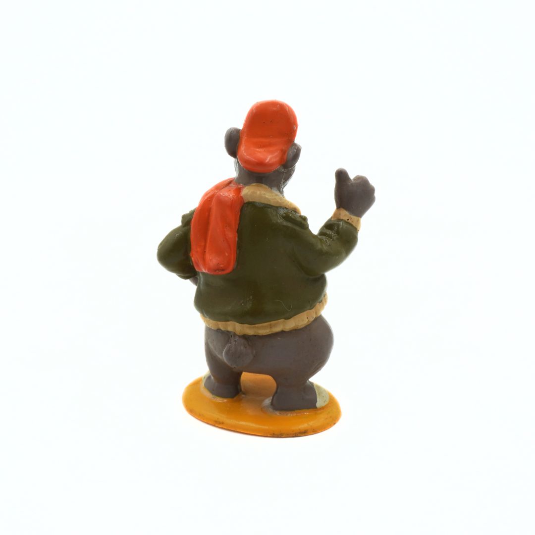 Small figurine of a person wearing a green jacket, red hat, and carrying a red bag on a white background