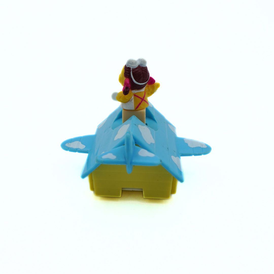 A small toy airplane house with a light blue wing and dark blue tail, a yellow body, and a small figure wearing a yellow outfit and brown hat standing on top, photographed on a plain white background.