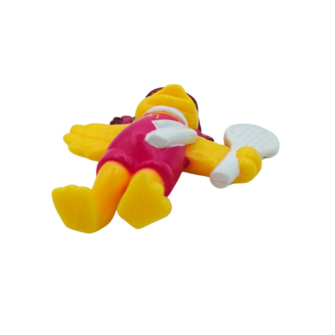 A small plastic toy figure of a cartoon character lying on its back, with yellow body parts, a pink outfit, and a white paddle-like accessory, photographed on a plain white background.