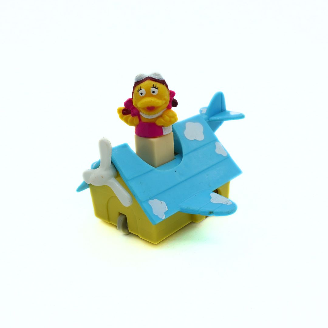 A vintage plastic toy airplane pop-up with a yellow base, light blue wings and tail, white clouds, and a small character figure on top, photographed on a white background.