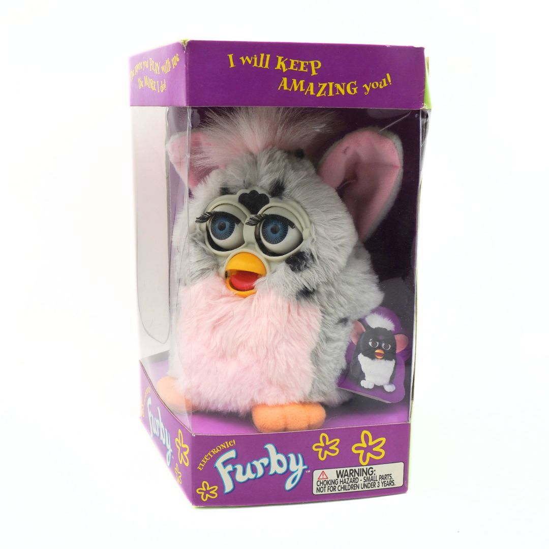 Plush toy in packaging with purple lid and base, featuring text and Furby branding.