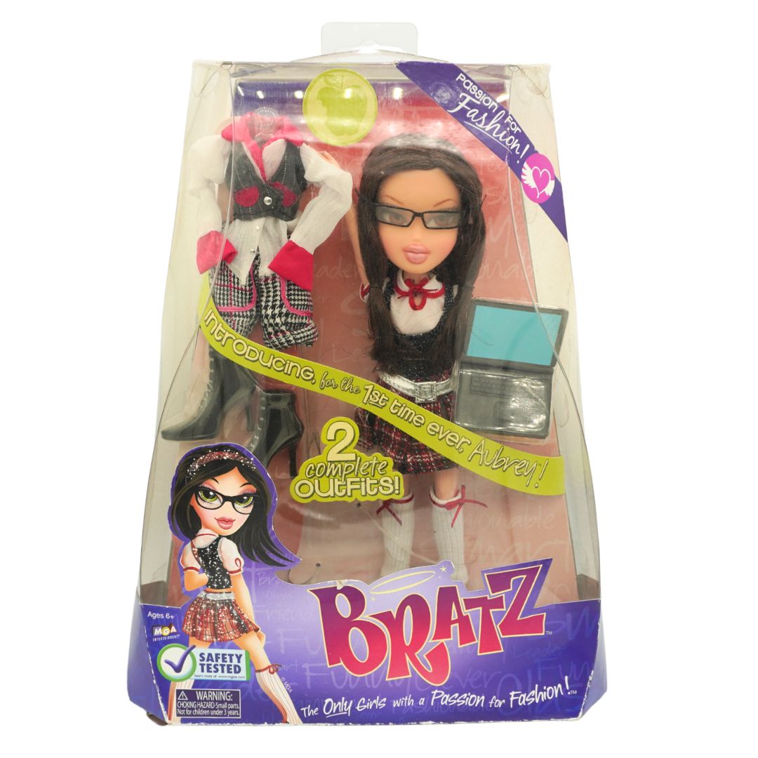 Bratz doll in retail packaging, featuring a dark-haired doll with glasses and a schoolgirl outfit, with an additional outfit visible behind her and a small teal accessory in the clear box.