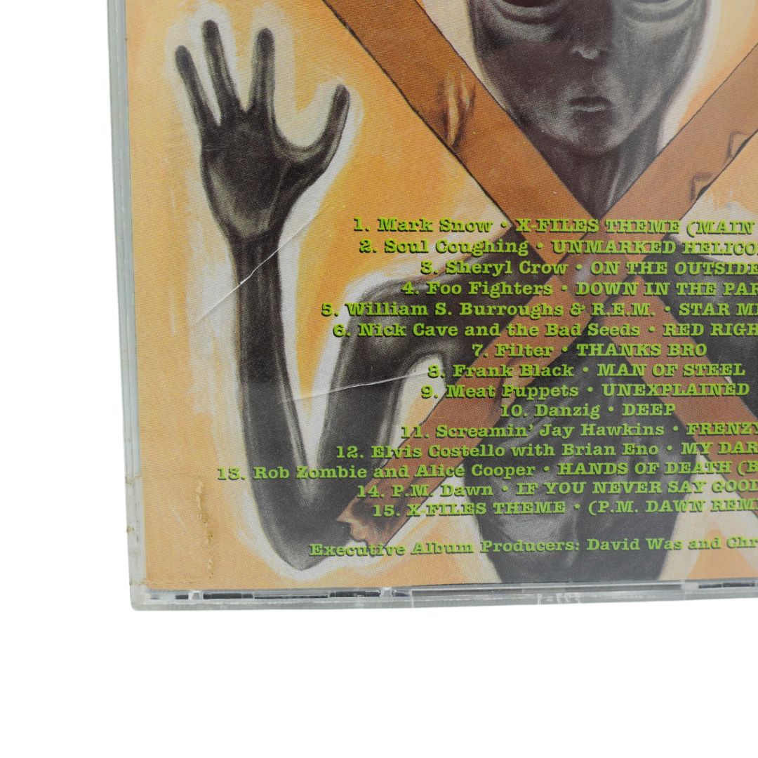 A used CD case showing the back cover of a music album, with a tan background, a black-and-white hand graphic, and green track listing text. The CD case is photographed on a white background.