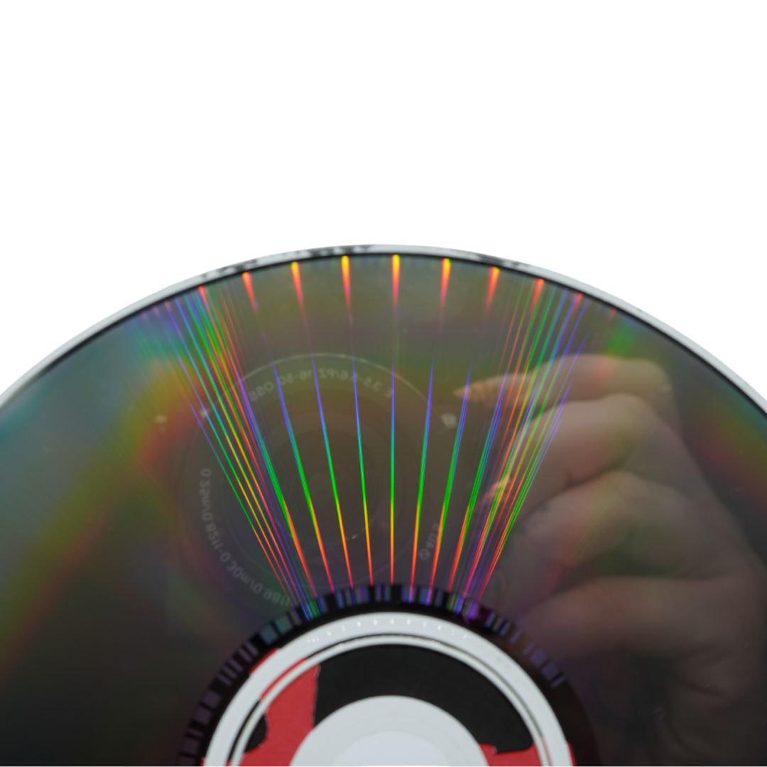 A close-up of a reflective CD or DVD disc showing rainbow iridescent light streaks, with part of a hand visible in the reflection against a white background.