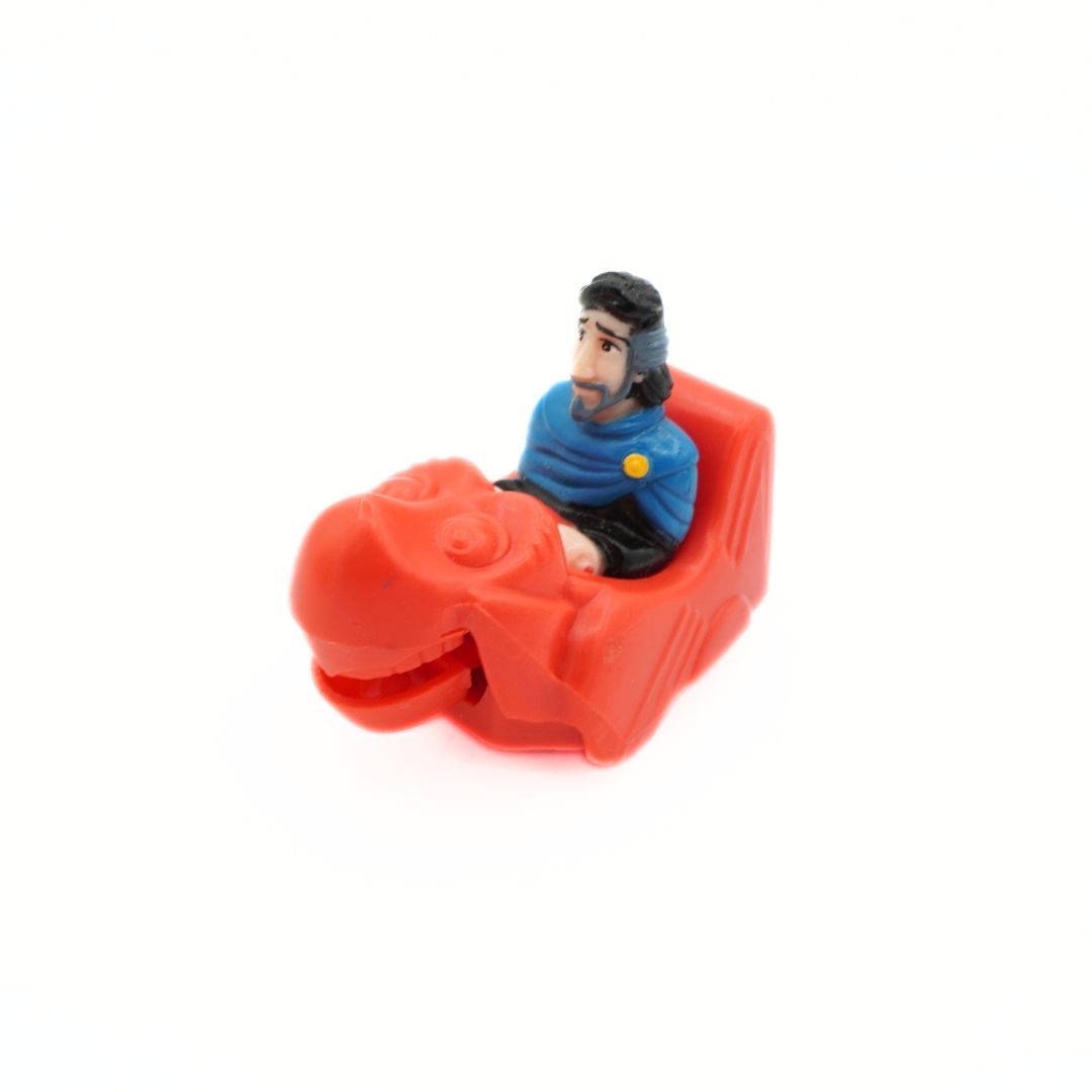 A small red dinosaur-shaped toy vehicle with a miniature figurine of a man in a blue uniform sitting inside, photographed on a plain white background.