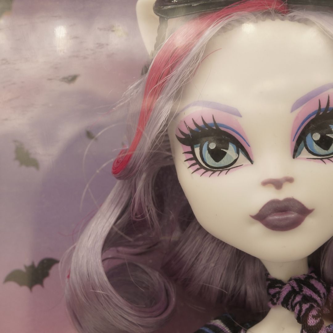 Close-up of a doll with purple hair and colorful makeup against a Halloween-themed background.