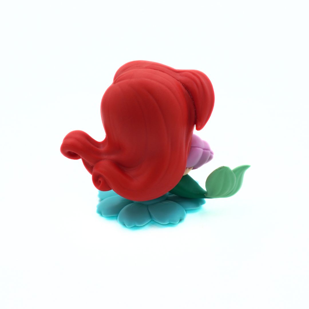 A stylized vinyl figure of Ariel from The Little Mermaid, viewed from behind, with her signature red hair and a flower base in shades of blue, green, and purple, set against a plain white background.