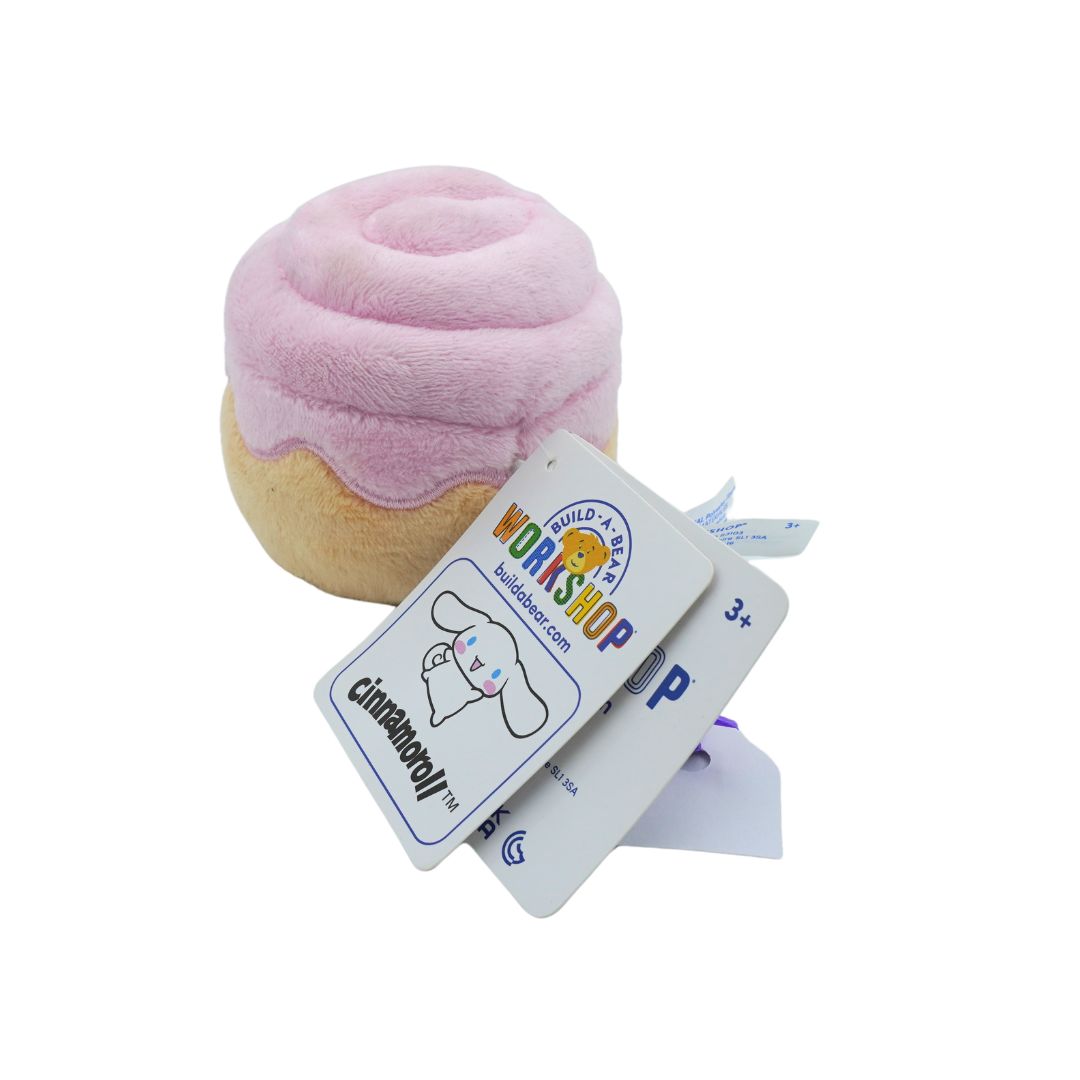 A plush cinnamon roll toy with pink frosting and a beige filling, shown with attached Build-A-Bear Cinnamoroll tags on a white background.