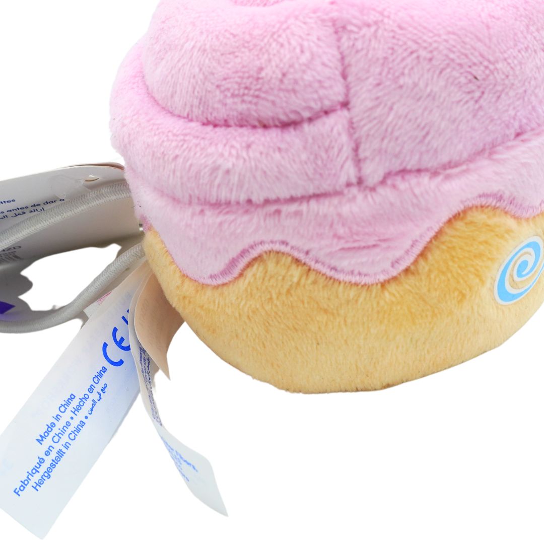 A plush toy shaped like a donut or cupcake with soft pink frosting-like top and tan base, shown close up on a white background with a tag and gray tool partially visible.