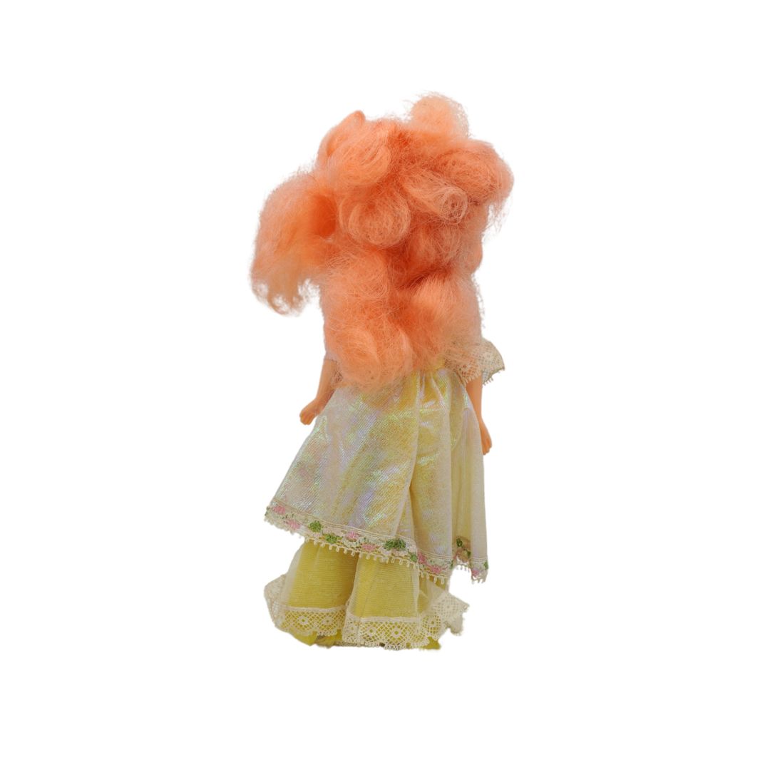 A doll viewed from the back, featuring curly peach-pink hair and a yellow dress with a white patterned overlay and lace trim, isolated on a white background.