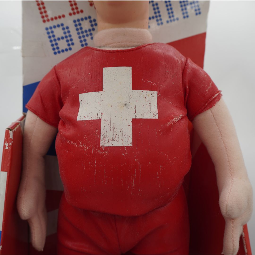 A vintage red doll or toy figure shown from the front, wearing a red shirt with a large white cross on the back, seated in a red-and-white box.