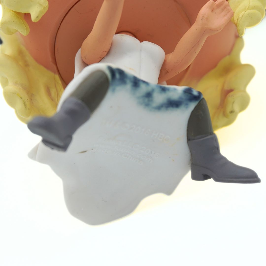 Close-up of a small figurine or doll lying on its back, wearing a white dress with blue shading and gray boots, against a plain white background.