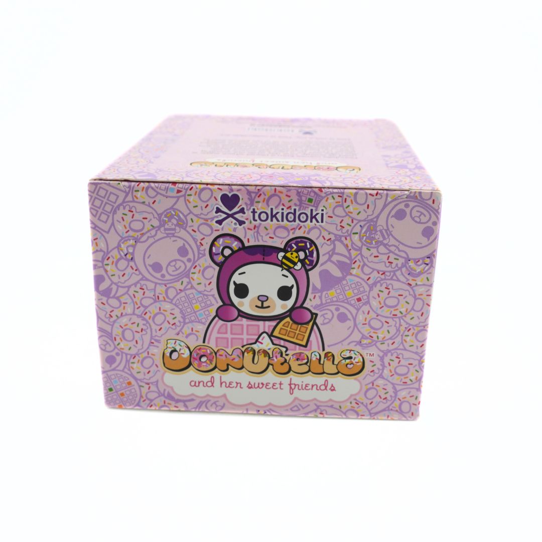 A square product box for 'tokidoki donutella and her sweet friends' featuring a cute panda character in a pink donut hat on a light purple background with donut patterns.