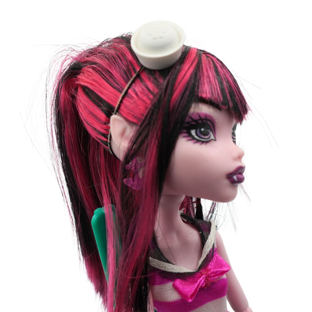 A close-up of a Monster High doll with long pink and black hair, a white hair clip, and a pink and beige top, shown against a plain white background.