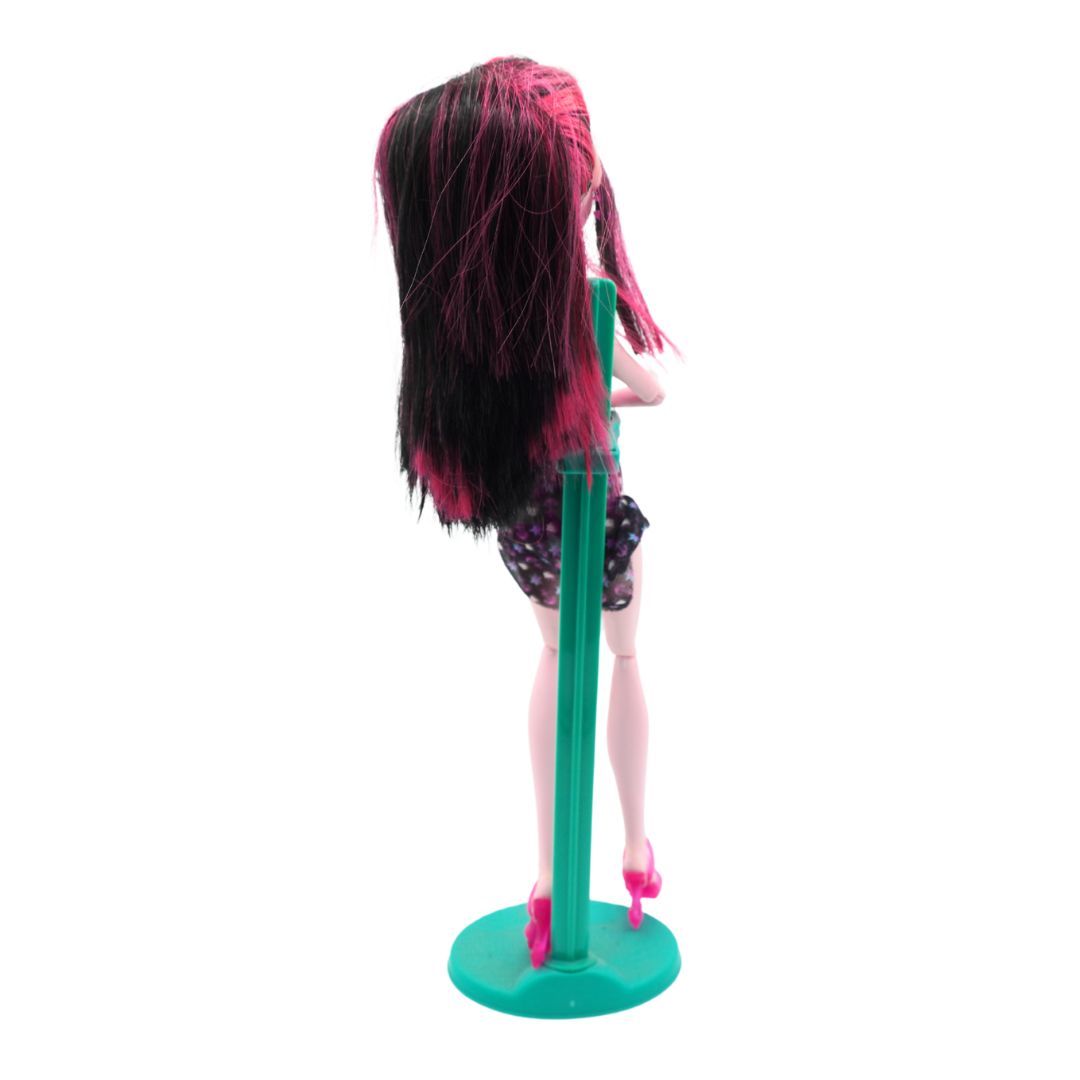 A Monster High doll shown from the back, featuring long black and hot pink hair, a black and pink polka-dot skirt, and pink high heels, standing on a green display stand against a white background.