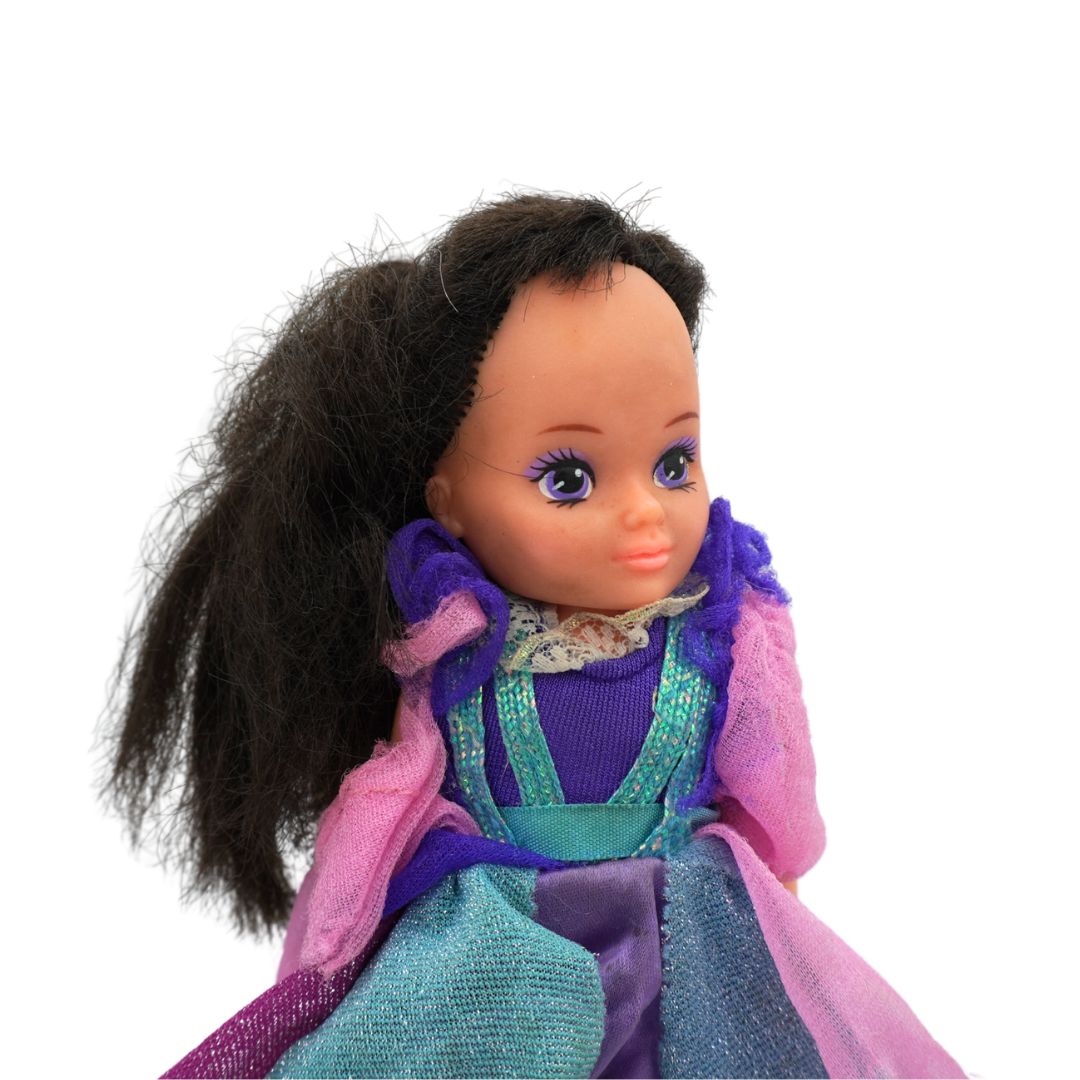 A vintage-style doll with dark brown hair, a purple and teal dress, and a pink tulle cape, shown against a plain white background.